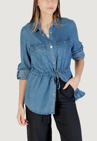 Camicia in denim con bottoni, maniche lunghe, vita regolabile con cordoncino, due tasche sul petto e texture morbida di colore azzurro chiaro.