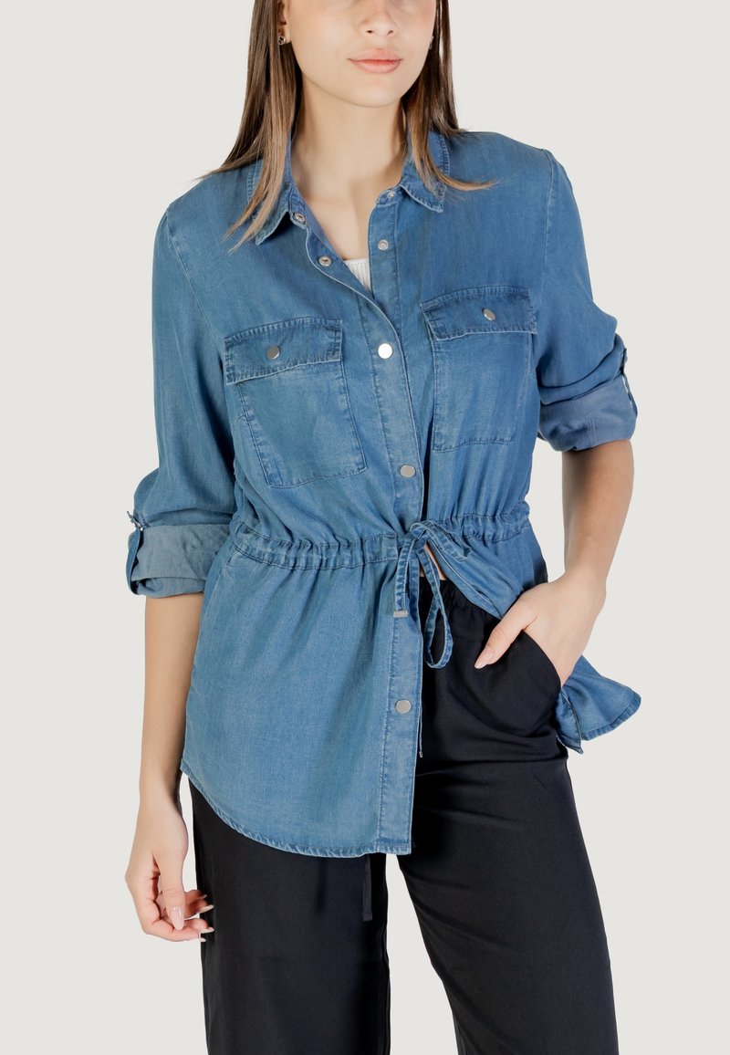 Camicia in denim con bottoni, maniche lunghe, vita regolabile con cordoncino, due tasche sul petto e texture morbida di colore azzurro chiaro.