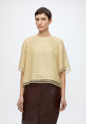 Γυναίκα που φοράει μπλούζα με υφή σε μπεζ χρώμα κοντομάνικη και φούστα από σκούρο καφέ δέρμα, στέκεται μπροστά σε απλό λευκό φόντο.