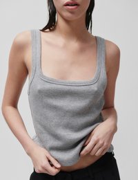 Νεαρό άτομο με βρεγμένα μαλλιά φοράει γκρι ριγωτό αμάνικο τοπ και μαύρο παντελόνι, προσαρμόζοντας το τελείωμα του τοπ με τα χέρια.