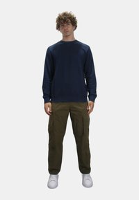 Felpa girocollo navy, pantaloni cargo oliva con tasche laterali e sneakers bianche. Outfit casual con una vestibilità morbida e un design semplice.