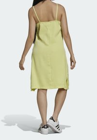 Kobieta w jasnożółtej, bezrękawnikowej sukience midi i białych butach Adidas z czarnymi paskami, stojąca tyłem do aparatu.