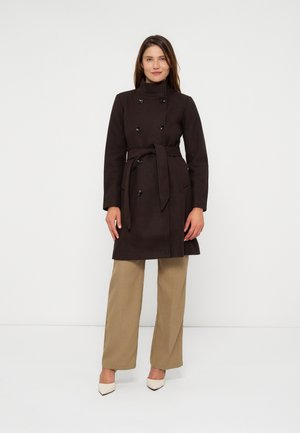 Femme portant un manteau croisé marron foncé ceinturé, un pantalon beige à jambes larges et des talons blancs pointus sur un fond blanc uni.