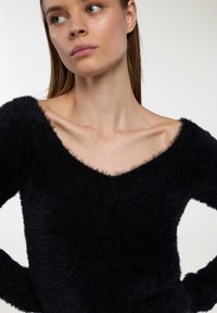 Maglione nero, peloso, con un design a scollo largo a V e spalline scoperte. La texture sembra morbida, con un bordo leggermente sfilacciato attorno al collo.