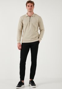 Maglione polo a maniche lunghe beige con colletto a mezza zip, abbinato a pantaloni neri slim-fit e sneakers nere con suola bianca.
