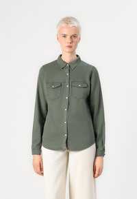 Camisa de ganga verde oliva com mangas longas, dois bolsos no peito, fecho de botão e gola mandarim. Textura suave; corte descontraído.