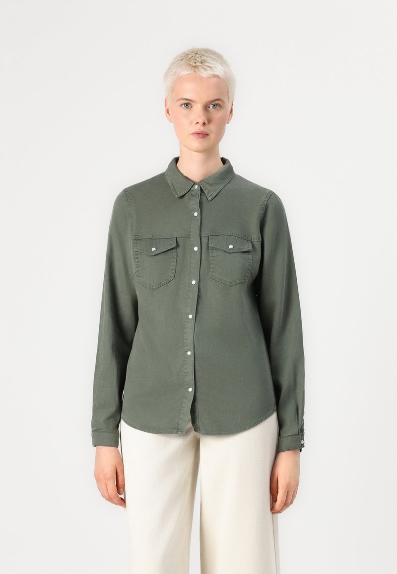 Camisa de ganga verde oliva com mangas longas, dois bolsos no peito, fecho de botão e gola mandarim. Textura suave; corte descontraído.