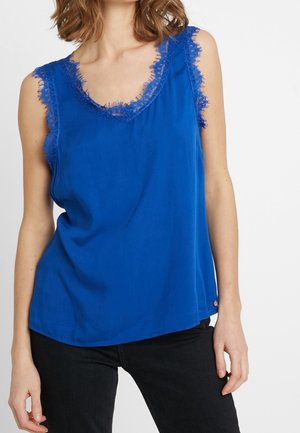 Kvinde iført en ærmeløs blå top med blondekant ved halsudskæringen og skuldrene, kombineret med sorte bukser.