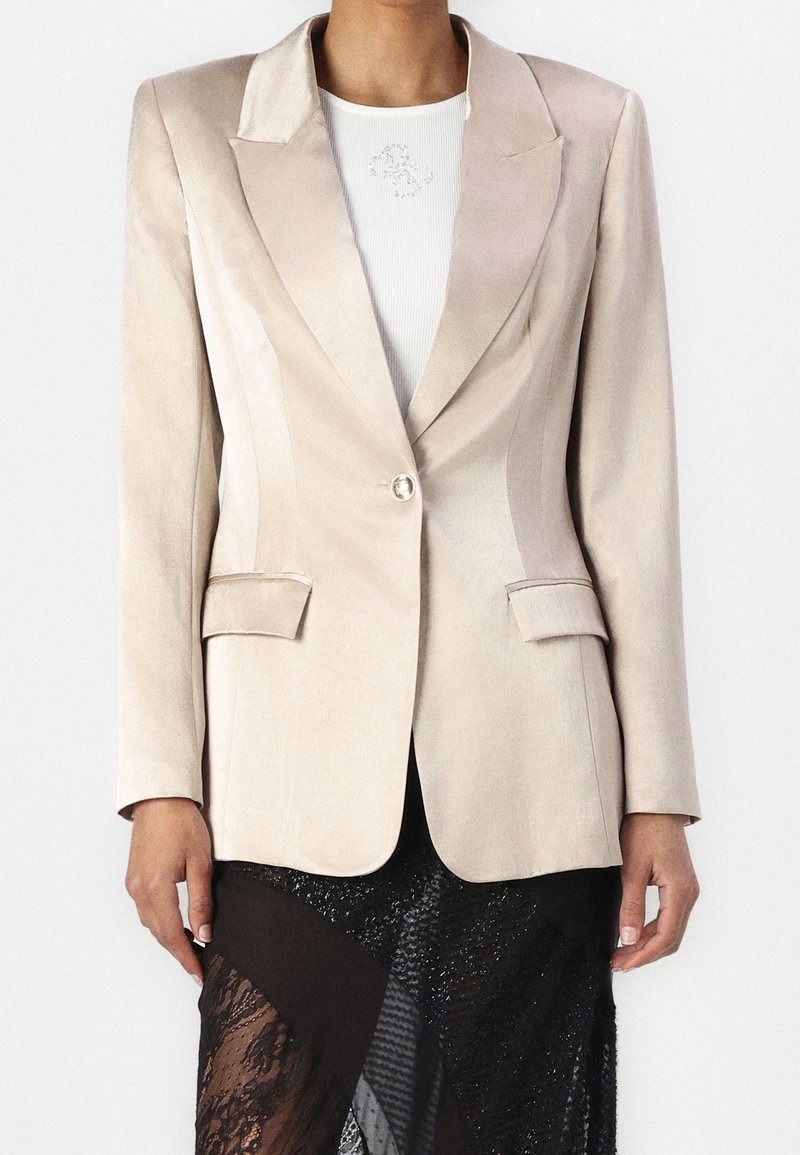 Femme portant un blazer beige à un bouton sur un haut blanc et une jupe en dentelle noire transparente avec des panneaux à motifs.
