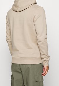 Sudadera beige con capucha ajustable, puños y dobladillo acanalados. Material suave, diseño entallado y textura lisa. Llevada sobre pantalones cargo oliva.