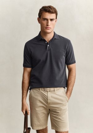 Giovane uomo con polo grigio scuro e pantaloncini beige, che tiene una borsa di pelle marrone con una mano in tasca, sfondo neutro.