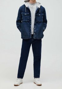 Jeansjacka med en grå huvtröja, två bröstfickor, mörkblå färg, avslappnad design, kombinerad med mörka jeans och vita sneakers.