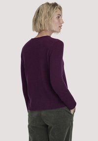 Blonde Frau mit kurzen Haaren steht seitlich, trägt einen lila Pullover mit langen Ärmeln und dunkelgrüne Cordhosen und hat die Hände in den Taschen.