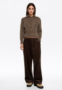 Mujer vestida con un suéter marrón de botones, pantalones de pana sueltos de color marrón oscuro y zapatos de senderismo color beige, de pie contra un fondo blanco.