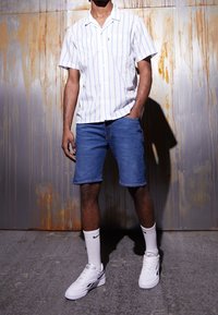 Camisa de botones con rayas blancas y azul claro, combinada con pantalones cortos de mezclilla. Lleva calcetines blancos y zapatillas deportivas con detalles en negro.