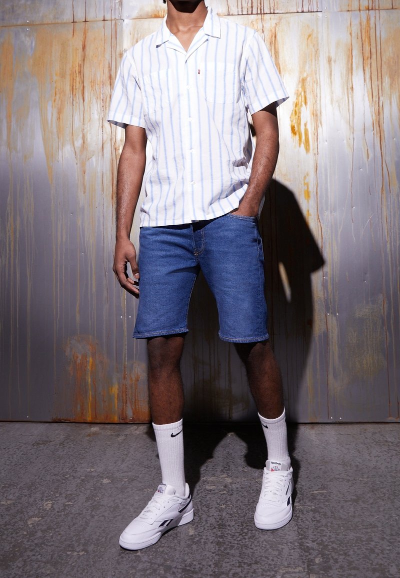 Camisa de botones con rayas blancas y azul claro, combinada con pantalones cortos de mezclilla. Lleva calcetines blancos y zapatillas deportivas con detalles en negro.