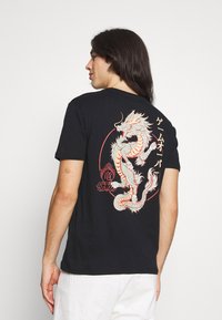 T-shirt de algodão preto com uma grande ilustração de dragão multicolorida nas costas, com texto estilizado e acentos circulares em vermelho e branco.