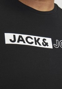 Crna pamučna majica s odvažnim bijelim natpisom "JACK& JONES" u različitim veličinama i stilovima fonta. Glatka tekstura, dizajn s okruglim izrezom.
