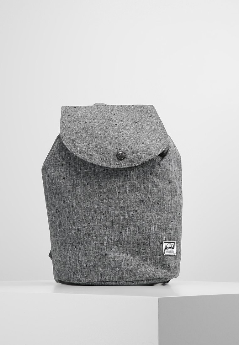 Sac à dos en tissu gris avec rabat et fermeture par bouton, petites taches noires et un écusson de marque rectangulaire en bas à droite à l'avant.