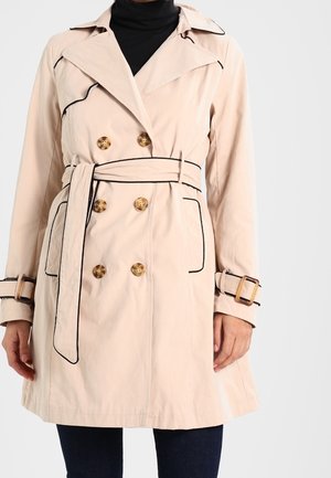 Beżowy dwurzędowy trencz z drewnianymi guzikami, czarną lamówką, paskiem i mankietami zapinanymi na klamry, noszony na czarnym golfie i ciemnych spodniach.