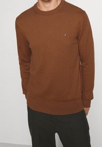 Homme portant un pull en maille brun avec un petit logo Tommy Hilfiger sur la poitrine, assorti à un pantalon foncé, sur un fond uni.