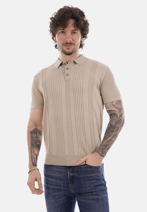 Uomo con capelli ricci che indossa una polo a maniche corte beige in maglia, jeans blu e tatuaggi visibili su entrambi gli avambracci, che posa davanti a uno sfondo bianco.