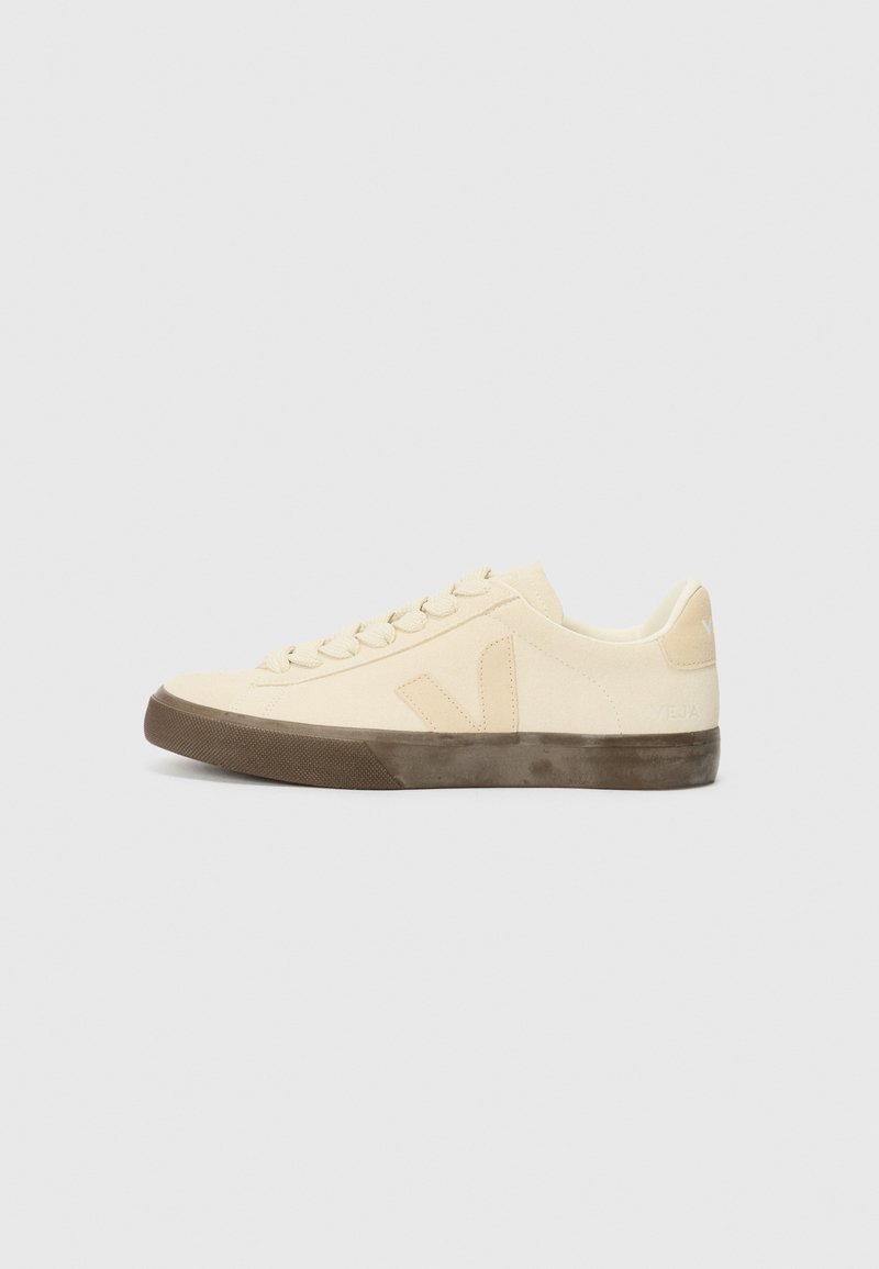 Beige canvas sneaker with round toe, flat laces, and textured brown rubber sole. Features a logo on the side and a minimalist design.