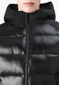 Doudoune noire avec capuche, dotée d'une finition brillante, d'un design matelassé et d'une fermeture éclair visible. Tissu texturé avec des accents lisses.