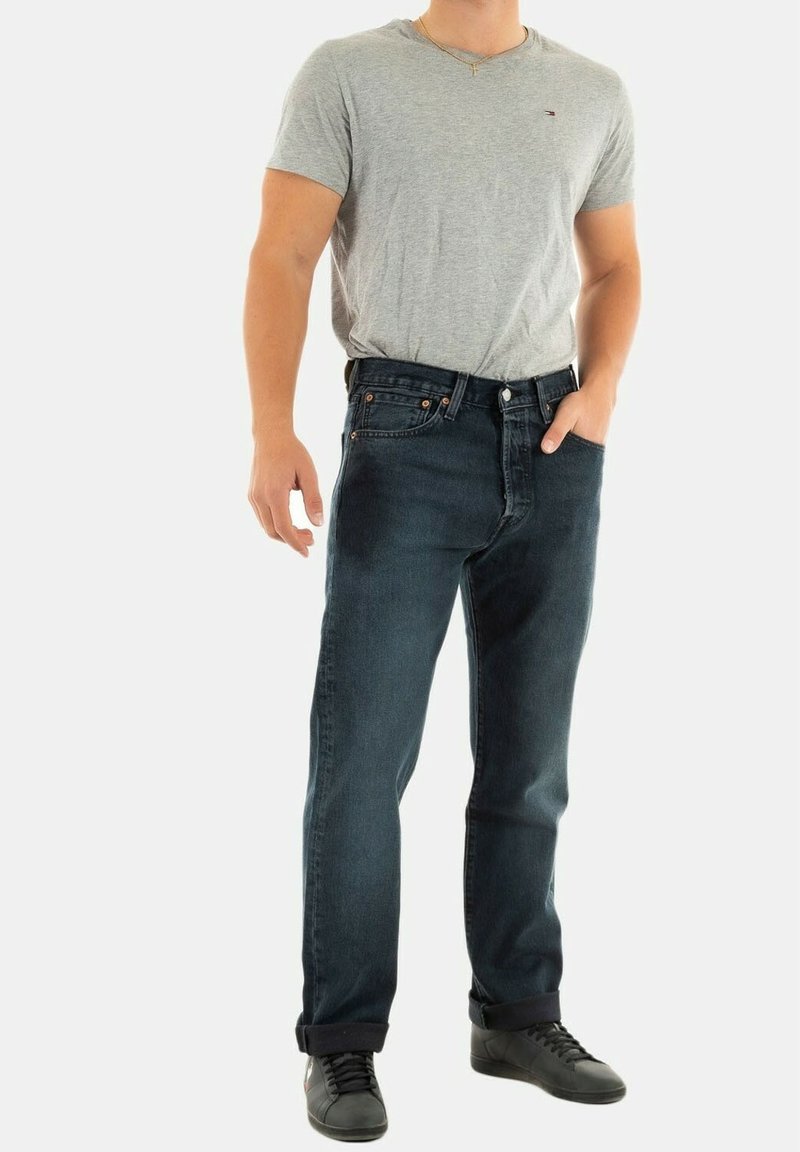 Jeans de mezclilla azul oscuro con un ajuste de pierna recta, combinados con una camiseta gris de manga corta y zapatillas negras. Costuras y bolsillos notables visibles.