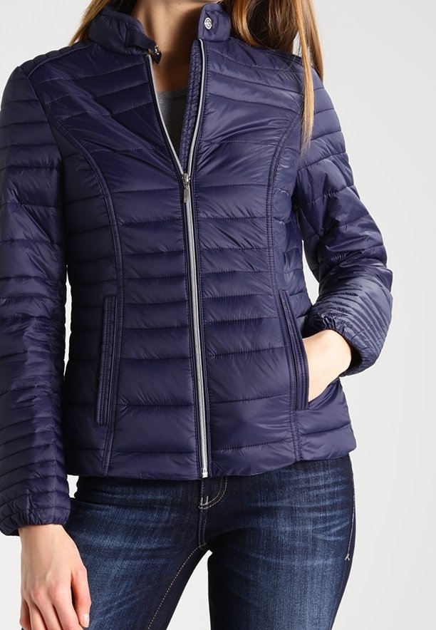 Femme portant une veste matelassée bleu marine ajustée avec une fermeture éclair à l'avant et des poches latérales, associée à un jean bleu foncé.