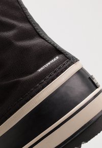 Bota impermeable negra con un upper texturizado y costuras reforzadas, suela de goma sólida y un contraste beige en el borde.