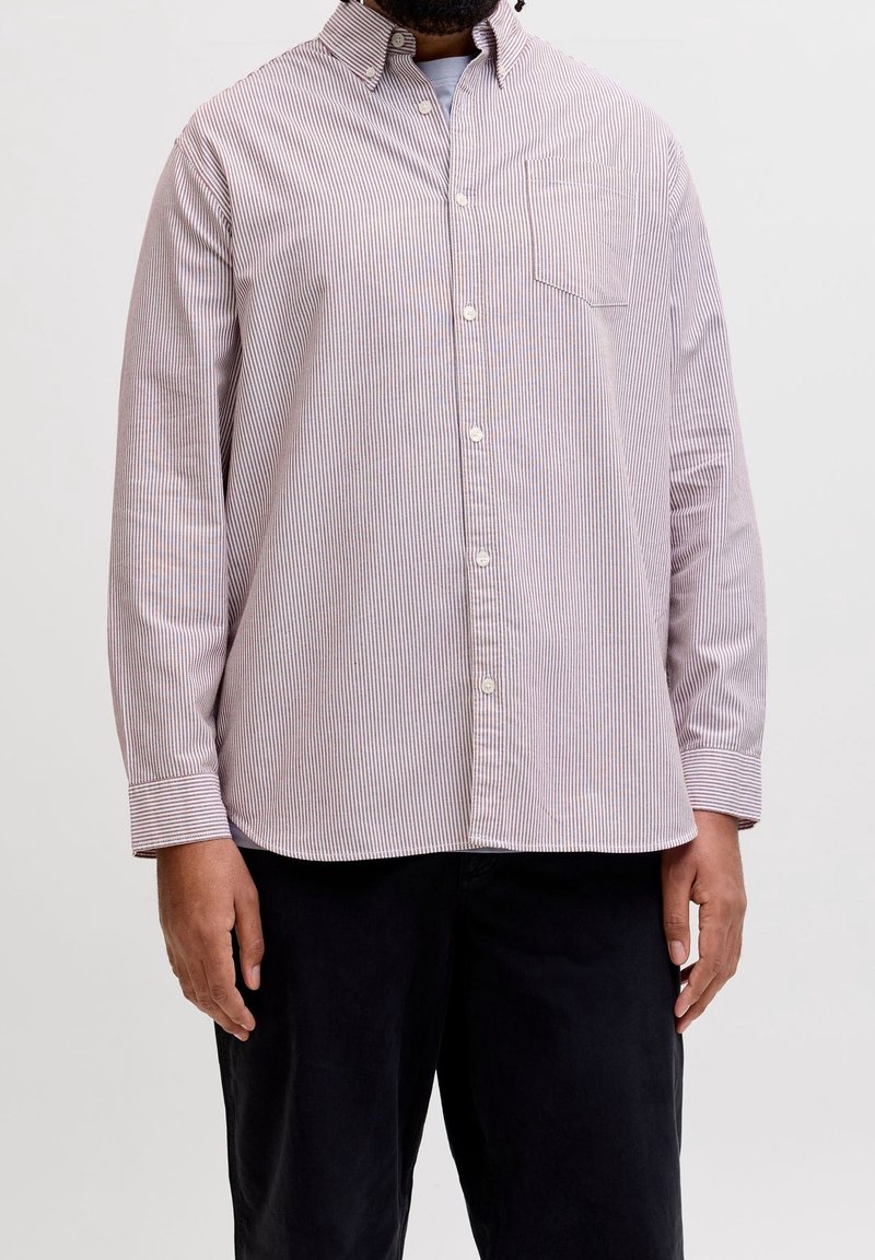 Homme portant une chemise à rayures claires à manches longues, boutonnée, avec une poche poitrine et un pantalon foncé sur un fond uni.