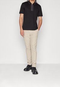 Černé polo tričko s límcem na zip, doplněné béžovými úzkými kalhotami a černými šněrovacími botami, na jednobarevném pozadí.