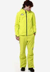 Giacca e pantaloni impermeabili di un brillante giallo neon, dotati di cappuccio, dettagli in cerniera nera e logo Kappa sul petto e sulla gamba dei pantaloni.