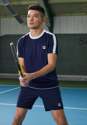 Marine sportshirt met witte accenten, logo op de borst, gecombineerd met bijpassende marineblauwe shorts. Speler houdt een tennisracket vast op een blauwe baan.