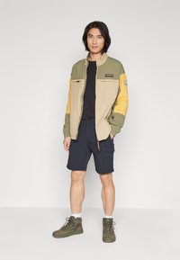 Giacca beige con maniche verdi e gialle, dotata di due tasche sul petto; abbinata a pantaloni corti blu navy e stivali alla caviglia verdi.