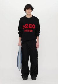 Czarny sweter z czerwonym napisem "032 BERLIN", w połączeniu z luźnymi czarnymi spodniami. Model trzyma niebieską dżinsową tkaninę, nosi czarne obuwie.