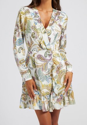 Kvinde iført en hvid langærmet kjole med farverige paisley-mønstre og et v-hals omslagsdesign, stående mod en ensfarvet baggrund.