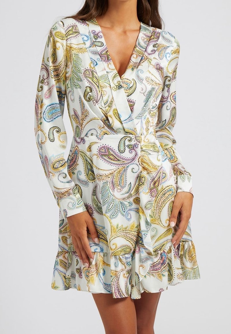Femme portant une robe blanche à manches longues avec des motifs cachemire colorés et un design portefeuille à col en V, debout devant un fond uni.