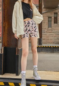 Jasno beżowa kurtka na czarnej koszulce z grafiką, połączona z wzorzystymi różowymi i czarnymi szortami oraz oliwkowymi trampkami za kostkę z białymi podeszwami.