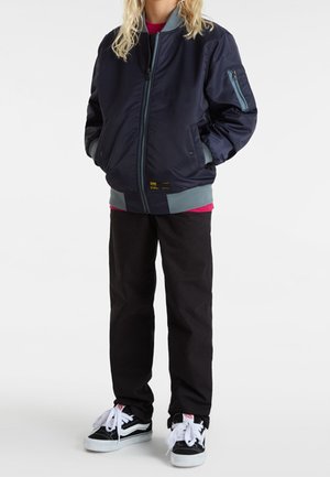 Barn iført en marineblå bomberjakke, sorte bukser, sorte og hvide sneakers, stående med hænderne i jakkelommerne mod en hvid baggrund.