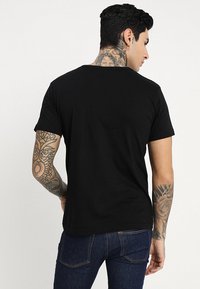 Schwarzes Baumwoll-T-Shirt mit Rundhalsausschnitt und kurzen Ärmeln, taillierte Passform, mit sichtbaren Tätowierungen an den Armen und am Hals.