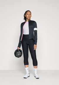 Zwarte sportjack met roze ritsdetails, gedragen over een lichtroze top en gecombineerd met zwarte leggings. Houdt een zwarte fietshelm vast.