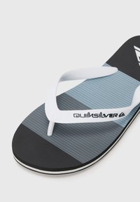 Ciabatte nere e grigie con superficie testurizzata, cinturino in gomma bianca e logo Quiksilver. Il design presenta blocchi di colore orizzontali.