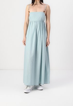 Vrouw draagt een mouwloze lichtblauwe maxi-jurk met dunne bandjes en witte plateau-sneakers, staand tegen een effen witte achtergrond.