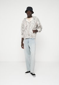 Grijze zip-up hoodie met bloemenpatroon, lichtbeige T-shirt, lichtblauwe straight-leg jeans en zwarte sneakers met witte zolen. Zwarte bucket hat.