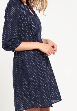 Frau trägt ein marineblaues Kleid mit weißen Tupfen und dreiviertel Ärmeln, seitlich vor weißem Hintergrund gezeigt.