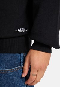 Black knitted sweater with a round neckline and wide cuffs, featuring a small white embroidered logo on the bottom left.