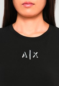 Naine kannab musta särki, mille keskosa all on särav hõbedane A|X logo, pilt on lõigatud ninast rinnani.