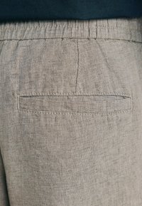 Gros plan sur un pantalon en lin beige avec une taille élastique et une poche passepoilée horizontale à l'arrière.